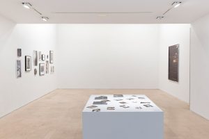 Gallery view of works on two opposing walls with a large white plinth in front.