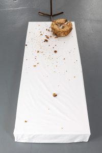 Low pedestal with a white table cloth and loaf of bread. The bread is torn apart and crumbs are everywhere.