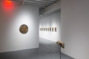 Gallery view showing the previous sculpture and a coin on the wall, as well as a series of framed works further down the hallway.