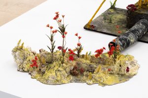 Closeup of flowers growing from the pile. A morphed or decomposing figure can be seen in the pile.