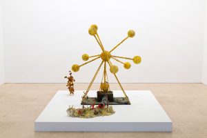 Straight on view of sculpture, large ggolden structure with small core wtih rods coming out, similar to an atom, with flowers in foreground.