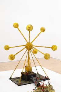 Large golden atom structure standing in the middle of a pedestal with a pipe coming out towards a collections of flowers growing from a patch of moss.