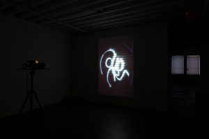 Gallery view of a projector located on the left of the image, beaming an abstract white mark to a wall in a dark exhibition space.