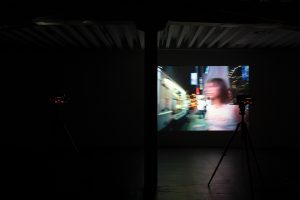 A photograph of a blurred figure walking in a urban nighttime street setting is projected onto a wall in a dark exhibition space.