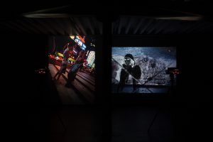 Two photographs of different orientations are projected onto a single wall in a dark exhibition space. The left image has a vertical orientation and is capturing a pair of women standing in the middle of Time Square, slouching their backs and with their hands held close to their faces. The right image is of an inverted figure overlaid on top of a close up image.