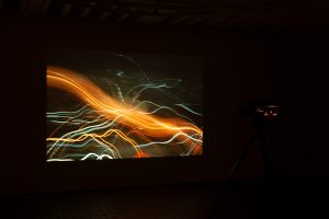 Gallery view of a photograph projected onto a wall in a dark exhibition space. The overexposed nighttime photograph captures a vividly moving multicolored light streak.