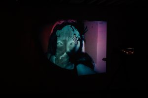 Gallery view of a photograph projected onto a wall in a dark exhibition space. The inverted photograph captures a medium close-up shot of a teal colored figure in a purple background as they stare intently at the camera.