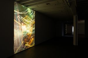 Gallery view of a photograph projected onto the left side wall of a dark exhibition space. The bombastic photograph captures multicolored light streaks with some running vertically across the composition while others spin around the image.