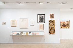 Gallery view of multiple artworks hung on the wall while printed material sits on a white shelf below the collection of hanged artworks.