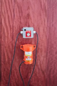 Gray and orange industrial vibrator with black wires affixed to red colored wood panel