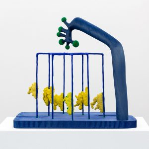 Another view of blue sculpture made of a blue base and thin blue rods with yellow sponge looking components attached to it. A thicker blue vertical form looms over the rods with green circular components attached to the top of base.
