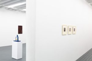 Gallery space with blue sculpture, brown painting, and three framed prints