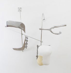 Nairy Baghramian's white plaster and aluminum sculpture named "Scruff of the Neck (UL 11, F)" is affixed to the gallery's white wall. Bulbous and organically shaped matte white plaster objects, partly coated with yellow bees-wax along the edges, are set in front of large uneven panes of cast and polished aluminum. These in turn are supported by dynamically mounted constructions of slender metal rods likewise polished to a silvery shine and affixed in two points to the gallery wall. Along the sides of several of these pieces of sculptural debris, hook-shaped proliferations reach out into thin air.