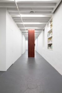 Flint Jamison's 2015 sculpture "Greaser" is a large scale red colored wood panel placed in the middle of a white gallery hallway with bookshelf on the right.