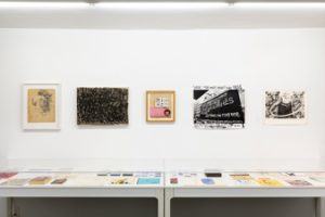 Gallery view of group exhibition "Doomed and Famous" in the basement gallery where there are five artworks hung on the white colored wall and multiple tables covered with glass below them that are displaying printed materials.