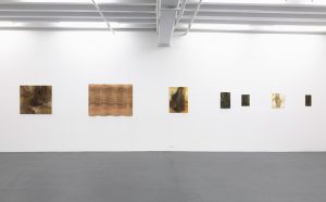 Gallery view of six paintings and a wooden carved panel on a white wall.