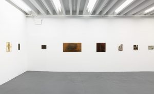 Gallery view of six paintings and two wooden carved panels across two white walls