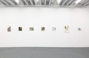 Gallery view of seven paintings arranged along a white wall.
