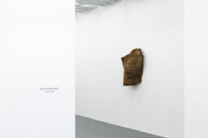 Gallery view of Beaux Mendes' Untitled redwood piece. It is a small, abstracted piece of redwood that hangs on a wall. To the left on the wall reads "Beaux Mendes Capitol Reef"